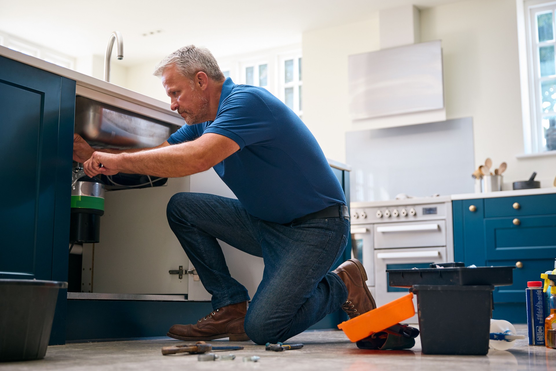 Mature Male Plumber Fixing Waste Disposal Unit In Domestic Kitchen Sink